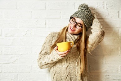 romantic dreaming hipster girl in knitted sweater and beanie hat with a mug in hands at white brick wall background. winter warming up concept.