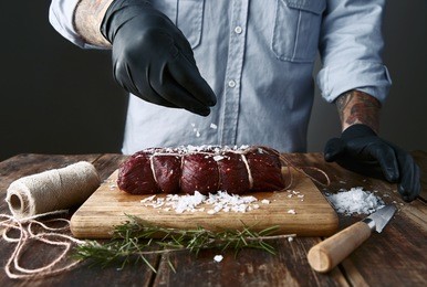 tattoed butcher in black gloves salts tied piece of meat to smoke it.