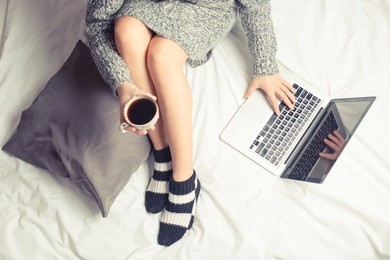 color picture of a beautiful young woman drinking coffee at home in her bed wearing a cozy sweater while checking her laptop