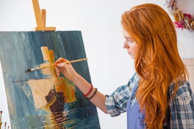 beautiful serene young female painter with long red hair painting picture on canvas in art workshop