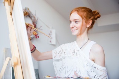smiling attractive young woman painter with red hair painting on canvas in artist workshop