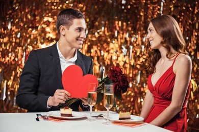 romantic photo of beautiful couple on glitter gold background. couple having date at valentine's day. lovers having dinner with champagne. young man giving valentine card and roses to his girlfriend