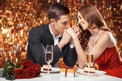 romantic photo of beautiful couple on glitter gold background. couple having date at valentine's day. lovers having dinner. there are glasses with champagne, desserts, roses and gift on table