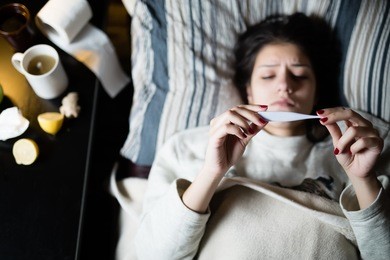 sick young woman in bed at home having flu,measuring temperature.thermometer to check temperature for fever.flu.virus.sick woman laying in bed.focus on thermometer