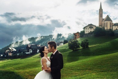 beautiful stylish asians bride and groom kissing in alpine meadows near the church in italy, trentino alto adige
