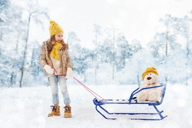 happy child girl plaing with a toy on a snowy winter walk. child rolls a teddy bear on a sled