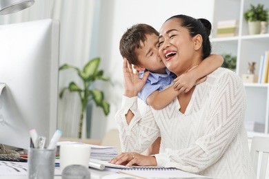 little indonesian boy kissing his mom, who is working in office