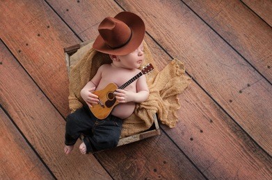 a three week old baby boy wearing a cowboy hat and jeans and playing a tiny acoustic guitar. he is lying in a wooden crate lined with burlap. shot in the studio on a rustic, wood background.