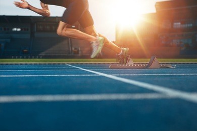athlete running on athletic racetrack. low section shot of male runner starting the sprint from the starting line with bright sunlight.