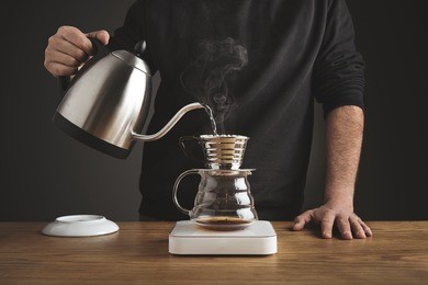 barista black sweatshirt spills hot water prepare filtered coffee from silver teapot to beautiful transparent chrome drip maker on white simple weights. everything thick wooden table cafe shop. steam