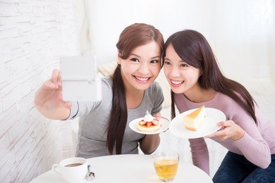 two happy young female friends take selfie picture by smart phone, with coffee cups and cakes in the living room at home, healthy lifestyle concept, asian beauty