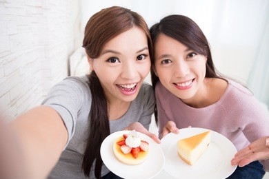 two happy young female friends take selfie picture with coffee cups and cakes in the living room at home, asian beauty