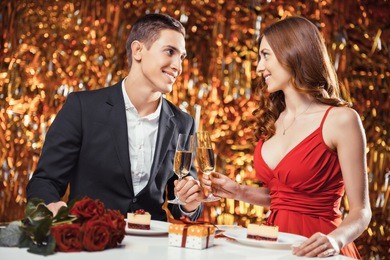 romantic photo of beautiful couple on glitter gold background. couple having date at valentine's day. lovers having dinner. there are glasses with champagne, desserts, roses and gift on table