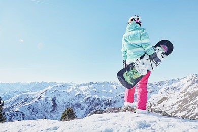 back view of female snowboarder wearing colorful helmet, blue jacket, grey gloves and pink pants standing with snowboard in one hand and enjoying alpine mountain landscape - snowboarding concept