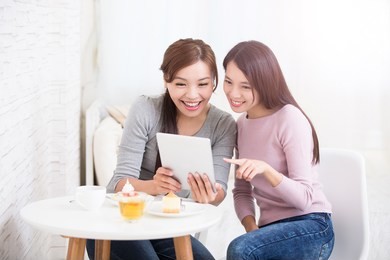 two happy young female friends use digital tablet pc with tea, coffee and cakes, healthy lifestyle and tecnology concept, asian beauty