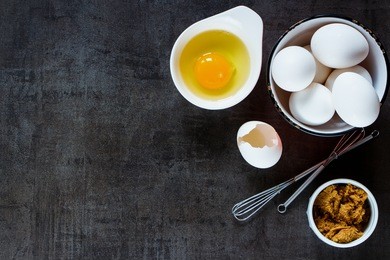 ingredients background: fresh eggs and brown sugar with whisk for baking on dark vintage table, top view, copy-space.