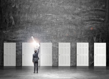 a young businessman standing in a room with numerous closed doors as if holding a magic key in a streched hand. back view. black background. concept of finding a way out.