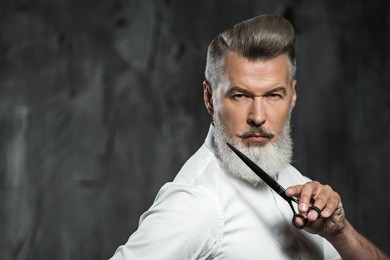 portrait of stylish professional hairdresser with beard. man wearing shirt, looking aside and holding scissors near his beard