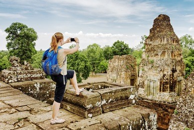 young female tourist with blue backpack taking picture with smartphone of the gopura from the top of ancient pre rup temple khmer in angkor on blue sky background. siem reap, cambodia.