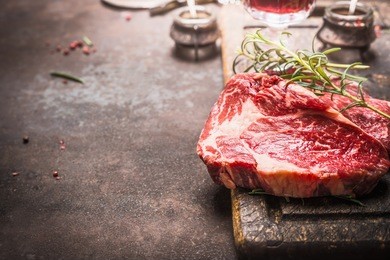 close up of raw fresh meat ribeye steak with herbs and spices on dark rustic metal background, place for text.