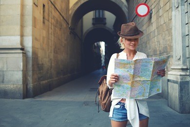 happy female tourist with backpack on shoulders exploring map while standing in alley near vintage building, trendy woman traveler with smile reading geographical atlas while touring in old city