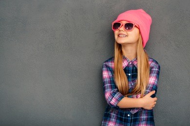 stylish and cute. cheerful little girl in sunglasses keeping arms crossed and looking up with smile while standing against grey background
