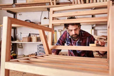 owner of an independent furniture design and manufacturing business, working on a wooden chair frame that he has made in his woodwork studio, carefully sanding it smooth