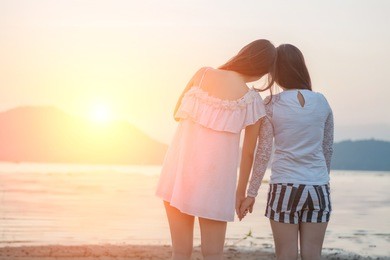 two young women hold hands together on the beach looking at sunset