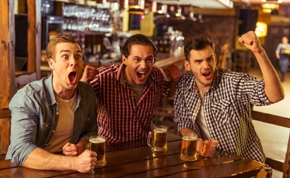three young men in casual clothes are cheering for football and holding glasses of beer while sitting in pub