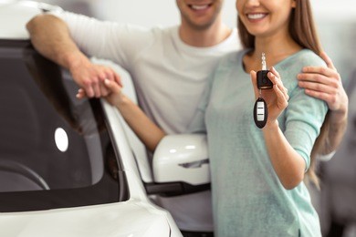 beautiful young couple is smiling and looking at camera while leaning on their new car in a motor show. woman is holding car keys, close-up