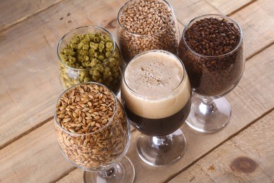 glasses filled with dark beer, different malts and hops over a wooden background