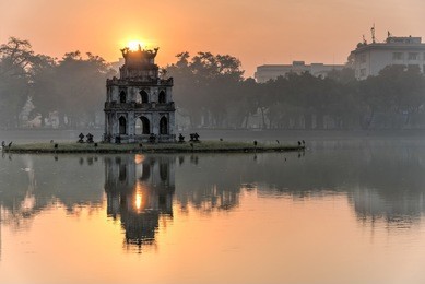 sun rising behind the turtle tower in the center of hoan kiem lake (lake of the returned sword). the lake is one of the major scenic spots in the city and serves as a focal point for its public life.