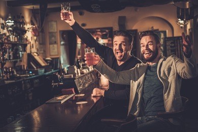 cheerful old friends having fun watching a football game on tv and drinking draft beer at bar counter in pub.