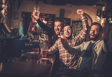 cheerful old friends having fun watching a football game on tv and drinking draft beer at bar counter in pub.