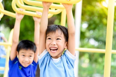 young asian boy hang the yellow bar by his hand to exercise at out door playground under the big tree.