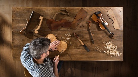 top view. craftsman luthier working on the creation of a violin. he sits at a wooden table in his workshop, various tools and instrument are placed on the workbench