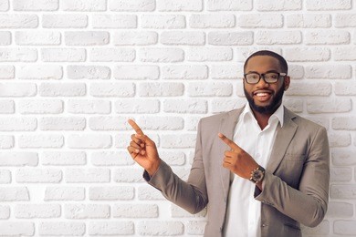 handsome afro american man in classic suit and glasses is smiling, looking at camera and pointing away, against white brick wall