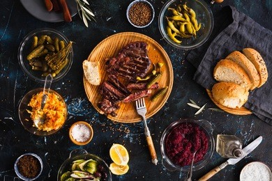succulent thick juicy portions of grilled fillet steak served with variety veggies dips, fermented veggies, cucumber and pepper marinated, bread buns on a old vintage table  authentic dinner party.