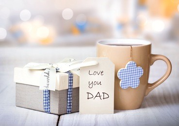father's day holiday greeting card.mug tea coffee and present gift box tag on wooden table blur lights background empty space.love you dad message.