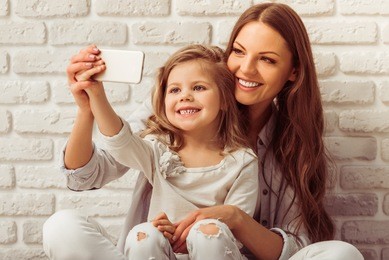 beautiful young mother is making a selfie with her cute little daughter using a smart phone and smiling, against white brick wall