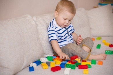 cute little toddler boy in a striped shirt playing with colorful plastic blocks on the sofa indoors. child having fun and building out of bright constructor bricks. early learning. creative.
