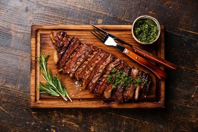sliced grilled beef barbecue striploin steak with chimichurri sauce on cutting board on dark wooden background