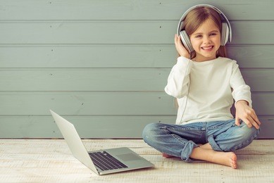 pretty little girl in headphones is listening to music using a laptop, looking at camera and smiling, against gray background