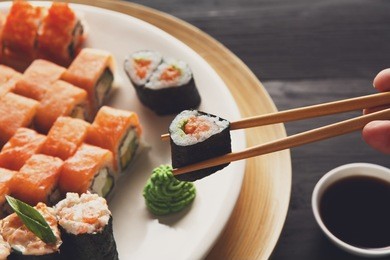 eating sushi rolls. japanese food restaurant, maki gunkan plate or platter set. closeup of hand with chopsticks taking roll. ginger, soy, wasabi dish at black rustic wood background.