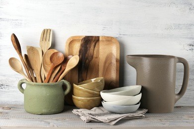 simple rustic kitchenware against white wooden wall: rough ceramic pot with wooden cooking utensil set, stacks of ceramic bowls, jug and wooden trays.