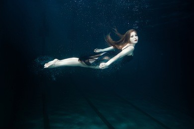 woman swims underwater with hands clasped, around the air bubbles.