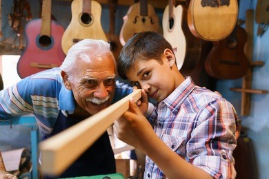 small family business and traditions: old grandpa with grandson in lute maker shop. the senior artisan teaches to the boy how to chisel wood to make a music instrument. 