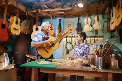 small family business and traditions: old grandpa with grandson in lute maker shop. the senior artisan gives teaches how to play classic guitar to the boy, who looks carefully at the instrument