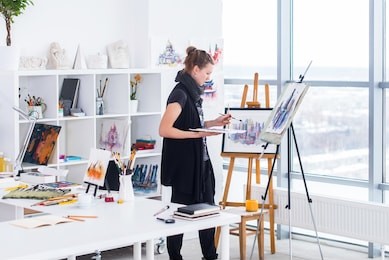 female painter drawing in art studio using easel. portrait of a young woman painting with aquarelle paints on white canvas, side view portrait