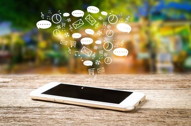 white smart phone on a wooden table emitting holographic image of social media related icons. blurred city background.